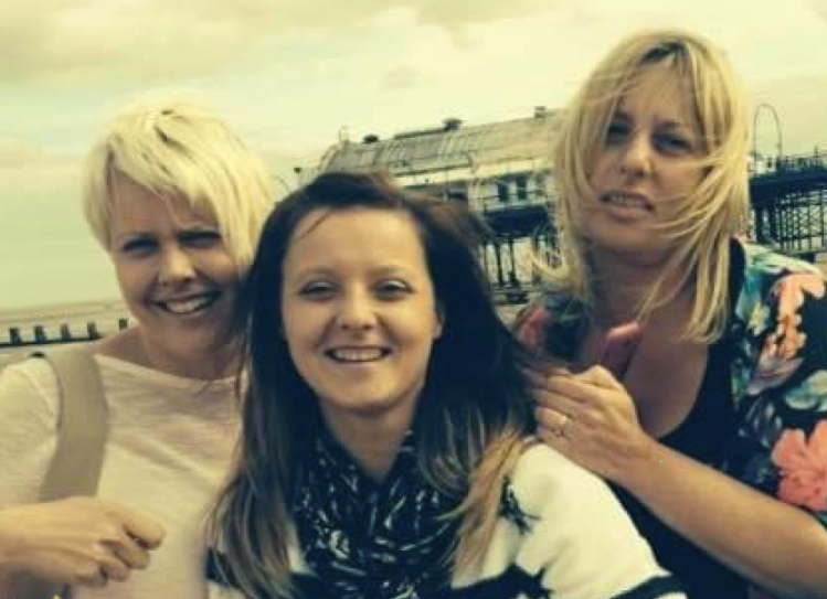 Lisa is smiling with one sister on her left and one on her right who are also smiling. There is a pier in the background.