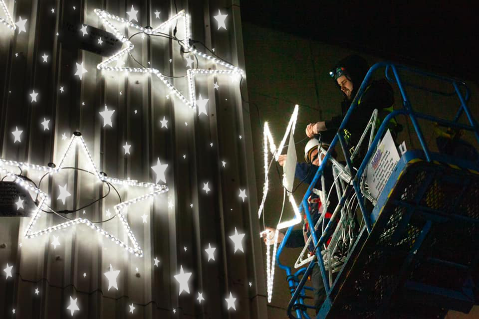 Two men are on a raised platform, placing large, illuminated stars onto the exterior of the hospital