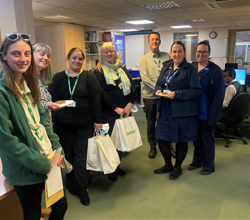 A group photograph of the Cancer Nurse Specialists in End of Life. Seven colleagues are standing in their office holding their Macmillan goodie bags. 