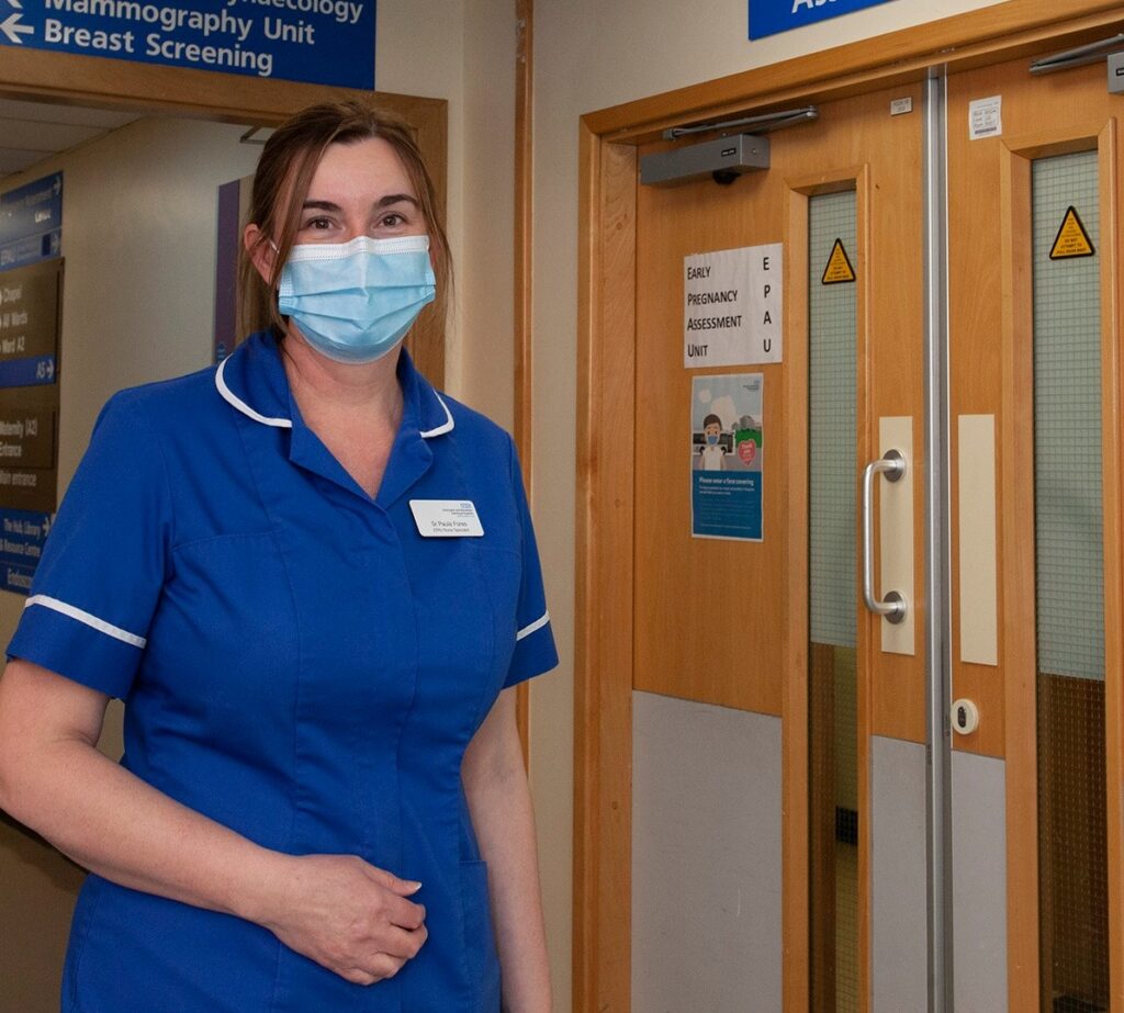 An photograph of Paula Fores, one of three new Early Pregnancy Loss Nurses at DBTH. She is a young, white woman with brown hair, wearing a blue nursing uniform and a mask. 