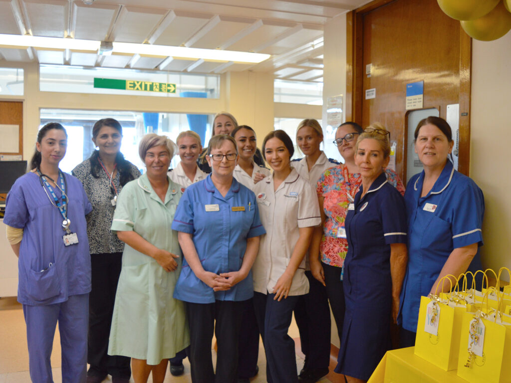 A group photograph of 12 colleagues from Ward 17. They are all female, dressed in blue and white clinical uniforms and scrubs., smiling at the camera in the ward corridor. 