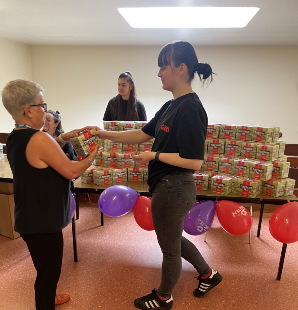 The volunteer is passing a box of tea to a staff member in front of a table filled with hundreds of boxes of tea and red and purple balloons 