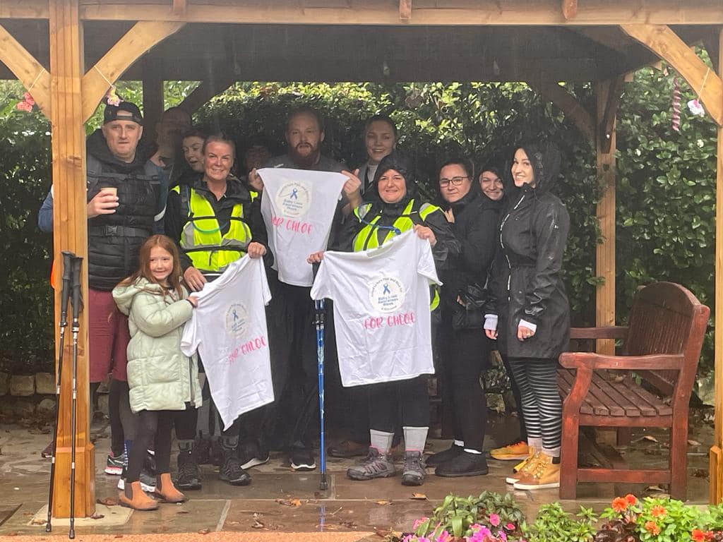 Mitchell and the group of walkers pose with t-shirts which say 'For Chloe' on them. They are all in walking gear, under a covered canopy from the rain