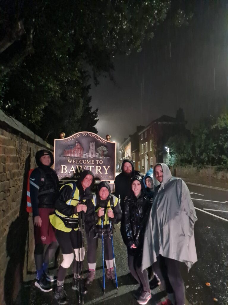 A group of people in walking gear and coats stand in front of the 'Welcome to Bawtry' sign, holding walking sticks. It is dark and raining heavily.