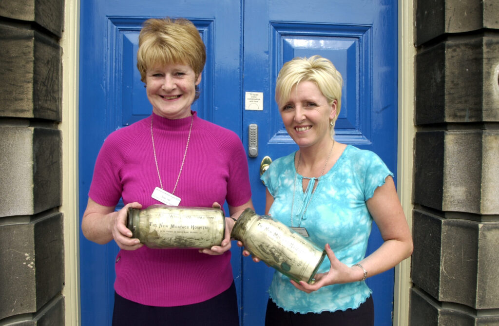Two female colleagues stood in front of a blue door holding glass jars out of the time capsule. 